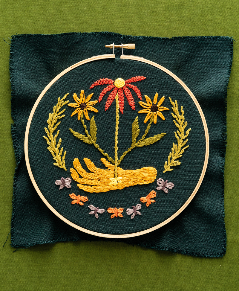 Embroidery by Christi Johnson. Yellow hand with flower blooming from it stitched on green fabric.